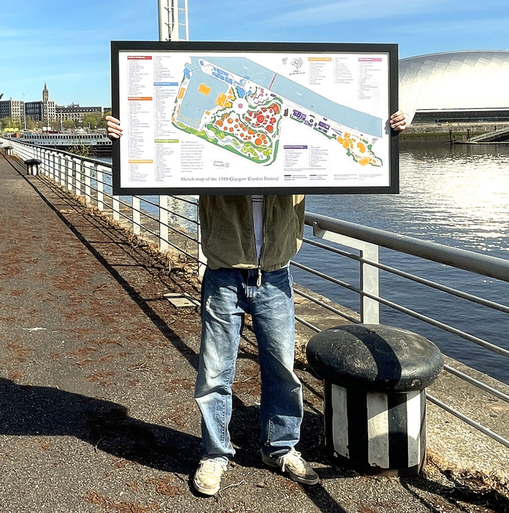 Image of a print of a sketch map of the 1988 Glasgow Garden Festival being held up at the location of the Festival